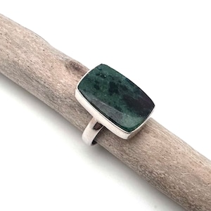 May include: A rectangular ring with a dark green stone set in a silver band. The stone has a mottled appearance. The ring is displayed on a piece of driftwood against a white background. The ring is a piece of jewelry.