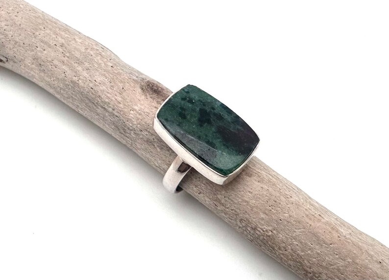 May include: A rectangular ring with a dark green stone set in a silver band. The stone has a mottled appearance. The ring is displayed on a piece of driftwood against a white background. The ring is a piece of jewelry.