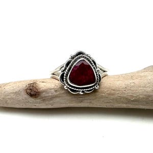 May include: A silver ring with a triangular red gemstone set in a decorative silver bezel. The ring is on a piece of driftwood.