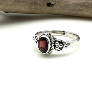 May include: A silver ring with a red gemstone set in a detailed bezel. The ring has a textured band and small decorative accents on either side of the stone.