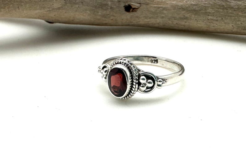 May include: A silver ring with a red gemstone set in a detailed bezel. The ring has a textured band and small decorative accents on either side of the stone.