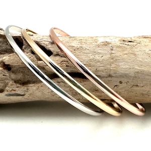Può includere: Tre braccialetti sottili in metallo, uno argento, uno oro e uno oro rosa, impilati su un pezzo di legno alla deriva.