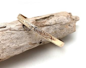Puede incluir: Un brazalete de oro con un diseño delicado e intrincado. El brazalete está hecho de un metal liso y pulido y tiene un aspecto simple y elegante.