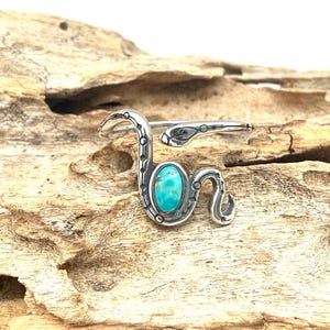 May include: A silver-toned snake ring with an oval turquoise stone. The ring is designed in the shape of a snake, with the head and tail wrapping around the finger. The ring is set against a natural wood background.