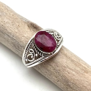 Op de afbeelding: Een zilveren ring met een ovaalvormige, dieprode robijn. De ring heeft een sierlijk ontwerp met wervelende patronen rond de robijn. De ring wordt gepresenteerd op een stuk drijfhout.