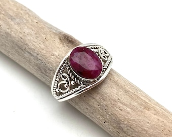 Puede incluir: Un anillo de plata con una gema de rubí rojo oscuro de forma ovalada. El anillo tiene un diseño ornamentado con patrones arremolinados alrededor del rubí. El anillo se muestra sobre un trozo de madera flotante.