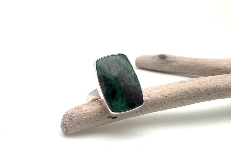 May include: A rectangular ring with a dark green stone set in a silver band. The stone has a marbled appearance with shades of green and black. The ring is displayed on a piece of driftwood against a white background.