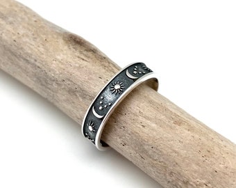 May include: A silver ring with a celestial design featuring suns, moons, and stars. The ring is set against a light-colored wooden background. The ring's band is decorated with detailed engravings of celestial bodies.