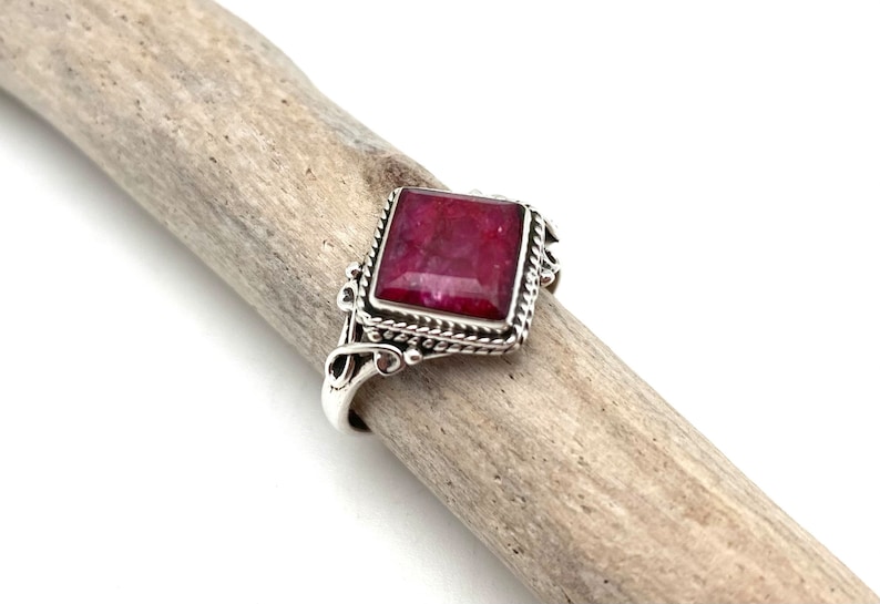 May include: A silver ring with a square-shaped red gemstone set in a decorative silver frame.