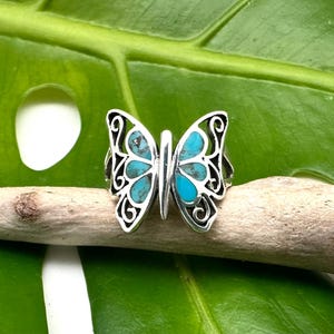 Peut inclure: Une bague en argent en forme de papillon avec des incrustations de turquoise. La bague est posée sur un fond de feuille verte.