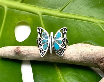 Op de afbeelding: Een zilveren vlinder ring met turquoise inleg. De ring ligt op een groene bladeren achtergrond.