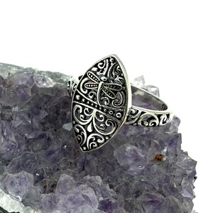 Könnte beinhalten: Ein silberner Ring mit einem ovalen, verzierten Gesicht mit einem Libellen-Design. Der Ring ist auf einer violetten Kristalloberfläche platziert. Die Libelle ist von aufwendigen Schnörkeln und dekorativen Details umgeben.
