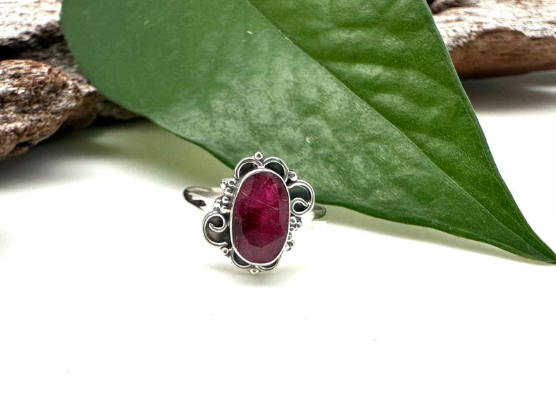 May include: A silver ring featuring an oval ruby gemstone set in a decorative filigree design.