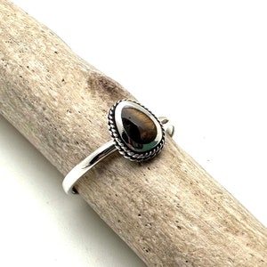 Könnte beinhalten: Ein Silberring mit einem tropfenförmigen Tigeraugenstein. Der braune Stein mit dunklen Streifen ist in einer gedrehten, seilartigen Lünette gefasst. Der Ring liegt auf einem Stück Treibholz.