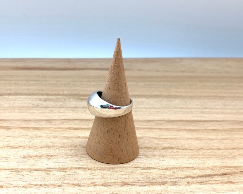 May include: A silver ring with a smooth, rounded design. The ring is displayed on a wooden ring holder.