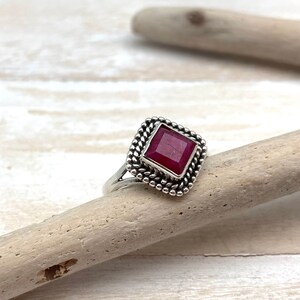 Op de afbeelding: Een zilveren ring met een vierkante robijn-edelsteen gezet in een decoratieve rand. De ring ligt op een lichtbruin houten oppervlak.