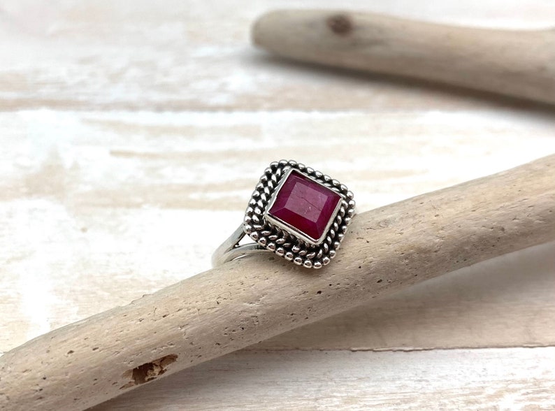 Op de afbeelding: Een zilveren ring met een vierkante robijn-edelsteen gezet in een decoratieve rand. De ring ligt op een lichtbruin houten oppervlak.