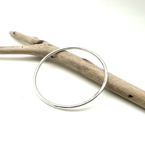 May include: A simple silver bangle bracelet, resting on a piece of driftwood.