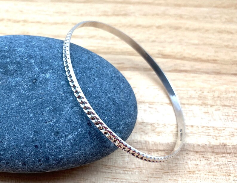 May include: A silver bangle bracelet with a textured design. The bracelet is on a gray stone surface.
