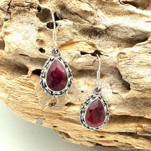 May include: A pair of sterling silver earrings with teardrop-shaped, faceted, deep red gemstones. The gemstones are set in ornate, openwork frames. The earrings hang from simple silver hooks, showcasing a classic jewelry design.