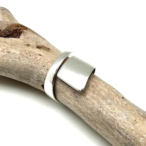 May include: A silver ring with a wide, flat band that wraps around the finger. The band is smooth and polished, and the ring is simple and elegant.