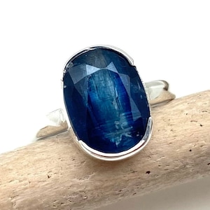 Peut inclure: Bague en argent ornée d'une grande pierre précieuse bleue ovale et facettée. La pierre est sertie dans un anneau en argent poli. La bague est présentée sur une surface en bois clair sur fond blanc.