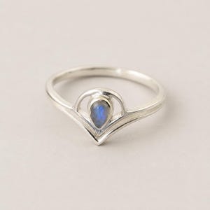 May include: A silver ring featuring a teardrop-shaped moonstone set within a V-shaped band. The metal has a polished finish, and the stone displays a subtle blue iridescence. The band curves upward to frame the central gemstone in a pointed setting.