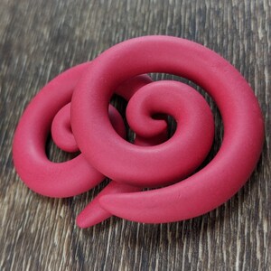 May include: A vibrant, raspberry-colored, spiral-shaped object rests on a wooden surface. The object appears to be made of a soft, pliable material, with a smooth texture. The design features two interconnected spirals, creating a unique, abstract form.