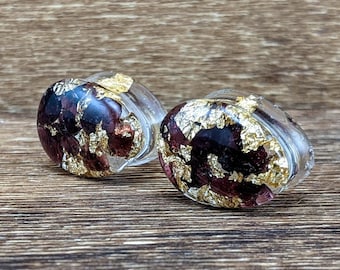 May include: A pair of clear resin earrings with a burgundy and gold leaf design. The earrings are oval shaped and have a shiny finish.