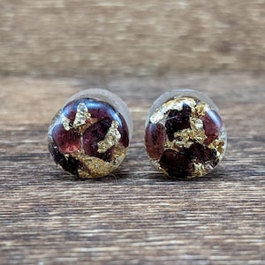 May include: A pair of round, burgundy resin earrings with gold leaf accents. The earrings are small and have a smooth, polished finish.