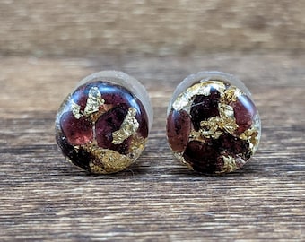 May include: A pair of round, burgundy resin earrings with gold leaf accents. The earrings are small and have a smooth, polished finish.