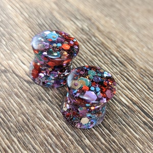May include: A pair of colorful glitter ear gauges. The gauges are made of clear resin with embedded glitter in shades of red, orange, blue, and purple.