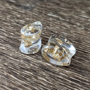 May include: Two clear acrylic ear gauges with gold flakes embedded inside. The gauges are shaped like a teardrop and have a slightly flared opening.