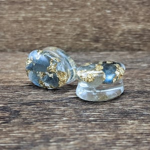May include: Two clear resin ear gauges with gold leaf inclusions. The gauges are round and have a smooth, polished finish.