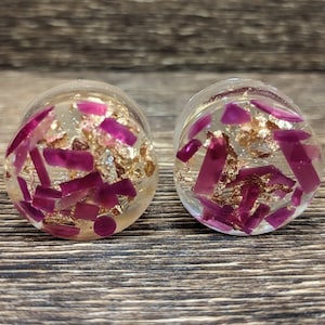 Puede incluir: Un par de medidores de oreja de acrílico transparente con purpurina dorada y rosa incrustada. Los medidores son redondos y tienen un acabado liso y pulido.