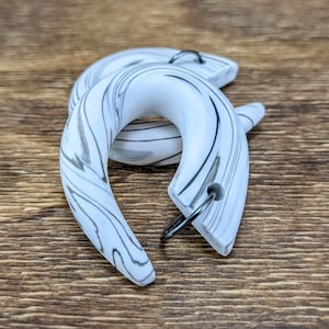 May include: A pair of white and gray marbled earrings with a curved, crescent shape. The earrings have a silver-colored metal hook. The earrings are displayed on a wooden surface. The text "SanguineCustom" is visible in the bottom left corner.