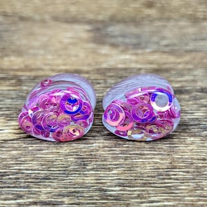 May include: A pair of teardrop-shaped ear plugs made of clear resin, each embedded with numerous small, iridescent pink and purple circular sequins that catch the light, creating a reflective and multi-dimensional appearance on the surface.