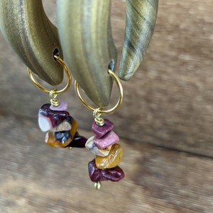 May include: A pair of gold-toned hoop earrings with a dangle of stacked, multi-colored, natural stone beads. The stones are shades of pink, red, brown, and yellow.