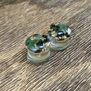 May include: Two clear glass ear gauges with green and gold flecks inside. The gauges are on a brown wooden surface.