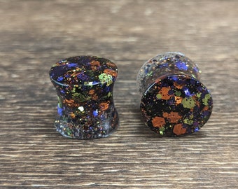 May include: A pair of black, orange, and purple glitter ear gauges. The gauges are made of resin and have a speckled appearance.