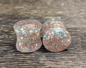 May include: A pair of iridescent pink glitter ear gauges. The gauges are made of resin and have a smooth, polished finish.