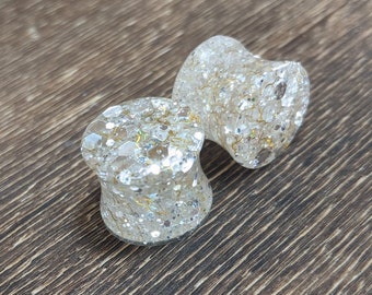 May include: Two clear, round, glitter-filled ear gauges. The gauges are on a brown wooden surface.