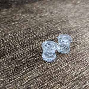 May include: Two clear glass ear gauges with a speckled, icy design. The gauges are on a brown wooden surface.