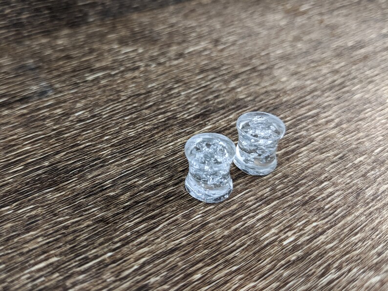 May include: Two clear glass ear gauges with a speckled, icy design. The gauges are on a brown wooden surface.