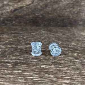 May include: Two clear glass ear gauges with a glittery, speckled effect. The gauges are on a brown wooden surface.