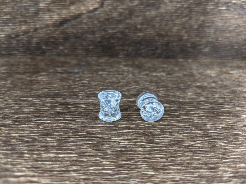 May include: Two clear glass ear gauges with a glittery, speckled effect. The gauges are on a brown wooden surface.