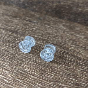 May include: Two clear glass ear gauges with a textured, cloudy appearance. The gauges are on a brown wooden surface.