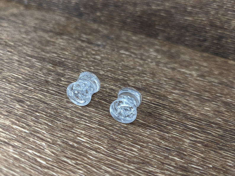 May include: Two clear glass ear gauges with a textured, cloudy appearance. The gauges are on a brown wooden surface.