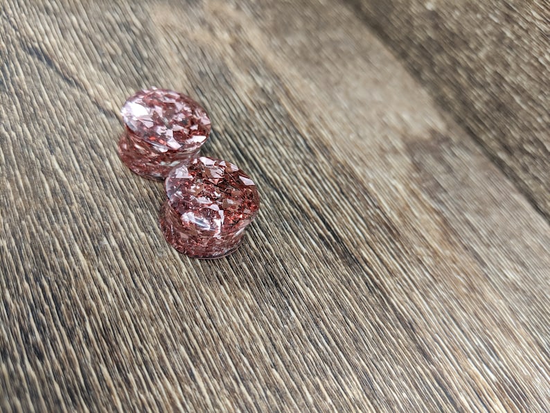 May include: Two round, clear, pink glitter ear gauges. The gauges are on a brown wooden surface.