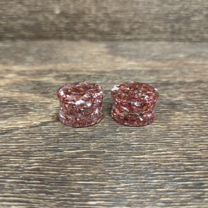 May include: Two round, red and gold glitter ear gauges. The gauges are made of resin and have a speckled appearance.